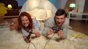 Front view laughing joyful couple gaming with game controllers looking at camera. Positive happy carefree Caucasian man and woman having fun enjoying hobby at tent in living room at home