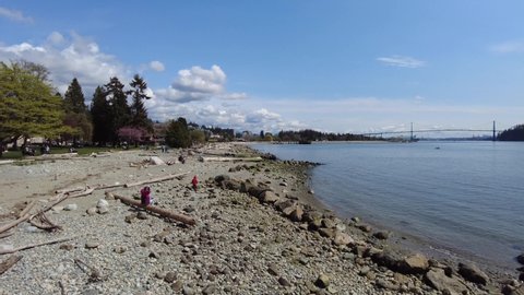 Vancouver, British Columbia, Canada - April 25, 2022: People sitting on a beach in North Vancouver seawall. Stanley Park and Lion's Gate bridge in the distance. 