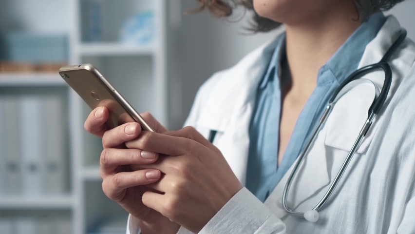 Female hands hold a smartphone phone with two hands writes a test sending a letter online mail. The girl doctor is in a medical office with a phone in her hands. Modern connection. Social media Royalty-Free Stock Footage #1089927413