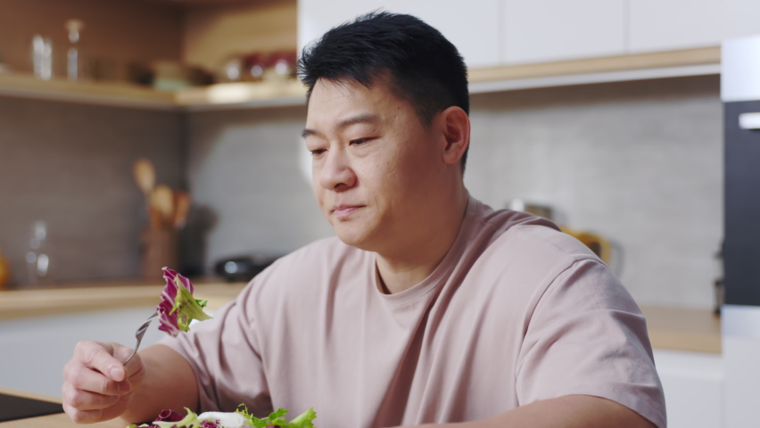 Portrait of sad Asian man sitting at table, holding fork in hand, trying to eat salad in cozy kitchen. Unhappy vegetarian guy. Facial expressions, healthy lifestyle. Indoors. Cooking. Diet concept. Royalty-Free Stock Footage #1089929501