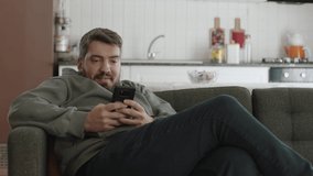 Young man looking at his smartphone while lying on the sofa is using smartphone, browsing social media accounts or chatting, texting or watching video.