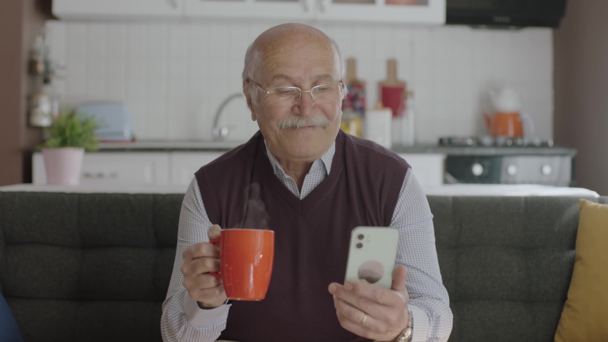 The old man dials the number on the smartphone. Old man sends sms to mobile phone while drinking coffee at home. 80s old man looking at mobile apps, sms and news on his smartphone. Royalty-Free Stock Footage #1089930763