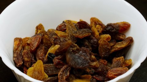 Brown raisins fall into a white plate. Light and dark raisins are poured into a bowl. Dried grapes falling on a white surface. Dried fruits. Healthy food. Flying food. Sweet dessert. Macro. Close-up