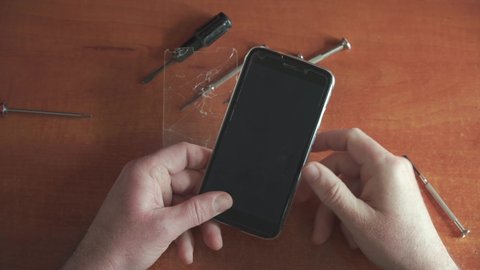 The repairman makes an inspection of the smartphone with broken glass. The protective glass on the phone is covered with cracks. Camera on top. Smartphone repair tools are on the table.