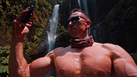 Athletic muscular man travel in rainforest jungle and making video on smartphone on background beautiful waterfall 4K Slow motion video