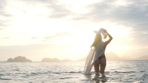 Wide Handheld Slow Motion Shot Of Young Woman In Thong And Sun Hat, Walking In Sea At Sunset, Corong Corong Beach, Philippines