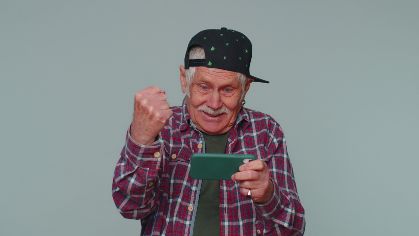 Worried funny addicted man in shirt enthusiastically playing racing video games on mobile phone. Elderly grandfather using smartphone gadget app with drive simulator isolayed on gray studio background