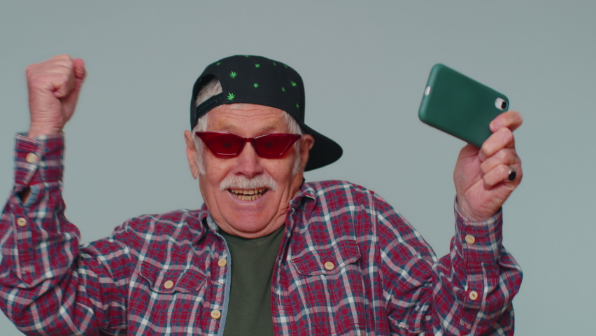 Worried funny addicted man in shirt enthusiastically playing racing video games on mobile phone. Senior grandfather using smartphone gadget app with drive simulator isolayed on gray studio background