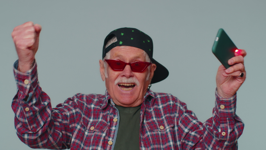 Worried funny addicted man in shirt enthusiastically playing racing video games on mobile phone. Mature grandfather using smartphone gadget app with drive simulator isolayed on gray studio background