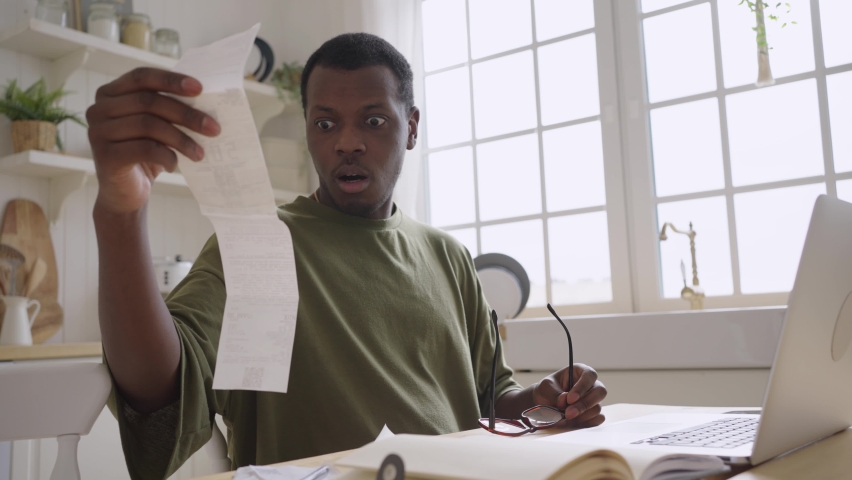 African American man looks at checks counting amount of money spent on shopping. Guy in glasses gets upset sitting in kitchen at home close view