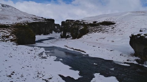 Stunning view of the entrance of famous gorge Fjaðrárgljúfur in southern Iceland near ring road with snow-covered rocks, steep cliffs and winding Fjaðrá river on sunny winter day with clouds.