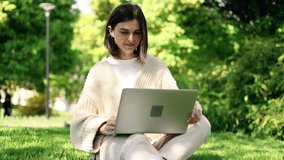 Dreamy attractive woman sitting on a grass in the park and talking on laptop by video chat. Student learning online.