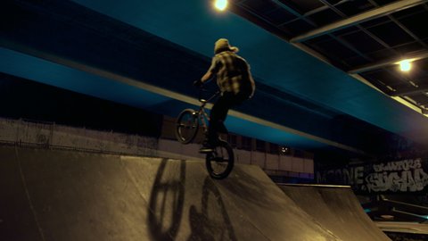 Young man practicing bmx bike in ramp at night urban skatepark. Casual bmx rider cyclist training jump trick stunt at skate park. Biker racing outdoors. Extreme sport youth culture recreation concept.