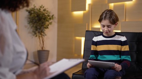 Boy patient looking at drawings on tablet and explaining what he sees to child counselor during psychological testing. Female child psychotherapist testing little patient during consultation