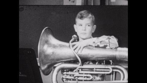 1950s: Boy holding tuba raises hand. Boy plays tuba. Teacher plays piano with one hand staccato style. Teacher adjusts electric setting and plays piano with both hands.