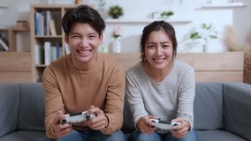 Happy millennia couple asian sitting on the couch playing video games, using joystick controllers. Woman and man in love have fun playing in video games console at home together.