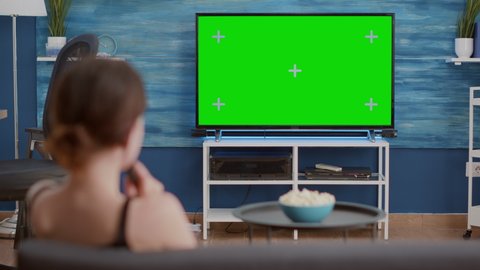 Static tripod shot of young woman switching channels while looking at green screen on tv and sitting on sofa. Over shoulder view of girl relaxing using television remote zapping on chroma key display.