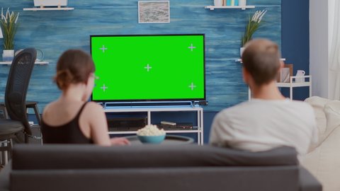 Couple sharing a bowl of popcorn while watching tv and switching channels looking at green screen in modern living room. Woman and man watching chroma key television and zapping while having a snack.