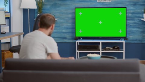 Sport fan sitting on sofa watching sport game on green screen tv mockup encouraging favourite team while eating popcorn. Man relaxing looking at television with chroma key display having a snack.