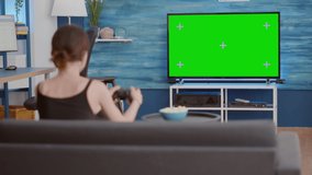 Static tripod shot of gamer girl holding wireless gamepad playing action console video game on green screen tv while sitting on sofa. Woman enjoying online gaming on chroma key display with controller