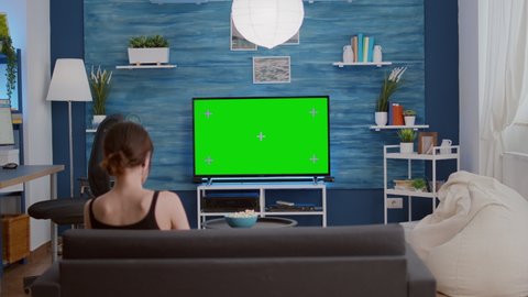 Young woman eating popcorn while relaxing watching movie on green screen television sitting comfortable on couch. Girl looking at tv favourite show on chroma key display in home living room.