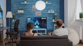 Boyfriend playing action space shooter console video game on tv while girlfriend is eating popcorn and giving him advice. Couple relaxing on sofa enjoying free time while gaming in modern living room.