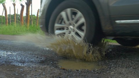 CLOSE UP: Car driving through road pit puddle and splashing water on paved road. Dangerous pothole with puddle in the middle of the asphalt road. Damage and bad road conditions in third world.