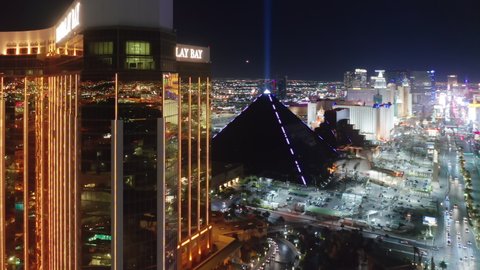 Las Vegas at night, Nevada, Apr. 2022. Breathtaking aerial amazing shiny golden building of Mandalay Bay luxury resort and incredible Luxor Pyramid hotel with Sphinx and bright beam into night sky 4K