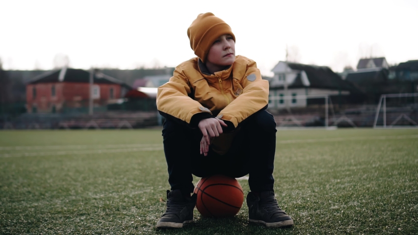 A boy is sitting on a ball on the grass at the stadium. He
