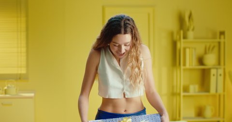 Slow motion of a portrait of a happy pretty young woman holding a gift box and feeling surprised. Close up of attractive model in sleeveless top having a present in studio with yellow interior.