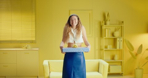 Happy satisfied smiling blonde woman in sleeveless clothes holding gift box and looking at the camera over yellow stylish interior at home. Joyful attractive young woman is holding present box