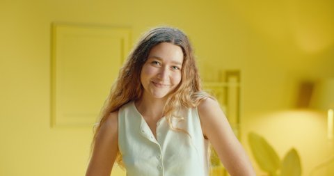 Slow motion of close up portrait of a pretty young brunette woman in sleeveless clothes smiling and looking at camera while standing in the studio with monochrome yellow color interior