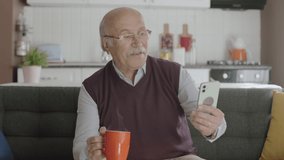 Old man watching funny video on his smartphone while drinking coffee at home. The man laughs at the sms or video he sees on his smartphone and looks at the camera.