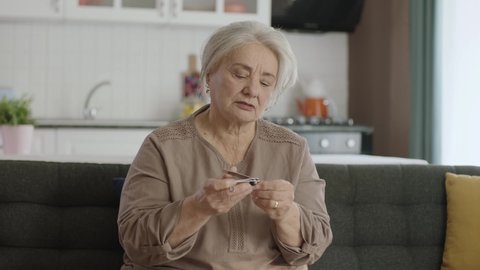 Elderly woman sitting in her armchair at home cutting her nails with nail clippers. Personal care concept.