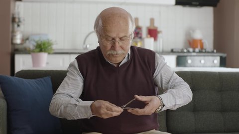 An elderly man sitting in his armchair at home is cutting his nails with nail clippers. The man accidentally cuts his nail and it hurts. Personal care concept.