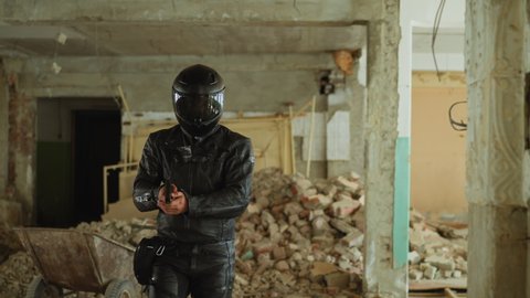 Motorcyclist in black jacket with helmet and handgun hides behind broken wall prowling about old abandoned building hall slow motion