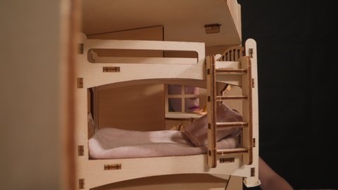 Little girl suffering from cold peeks through window of miniature wooden house with doll at table and bunk bed in dark room closeup zoom in
