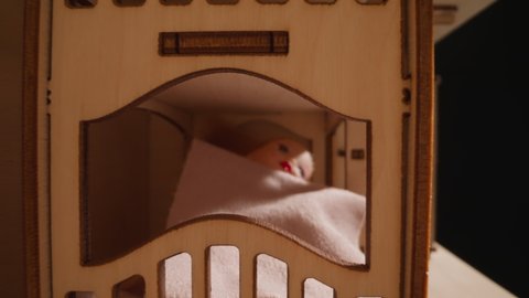 Cute doll lies under fleece fabric comforter on toy two-level wooden bed in dollhouse while sun rises up and new day begins closeup zoom out
