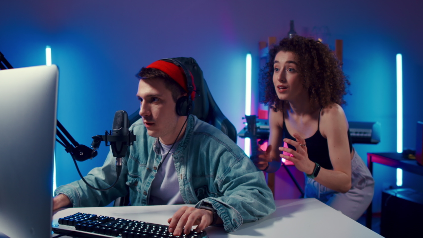 Excited Gamer Guy and Young Woman wearing headphones playing computer game neon fashion room, winner. Gamer winning hard match, guy's girlfriend is happy and congratulates her boyfriend looking at Royalty-Free Stock Footage #1089972709