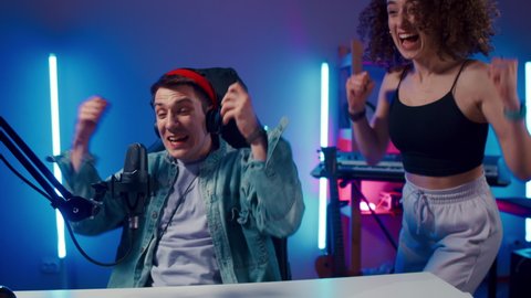 Excited Gamer Guy and Young Woman wearing headphones playing computer game neon fashion room, winner. Gamer winning hard match, guy's girlfriend is happy and congratulates her boyfriend looking at
