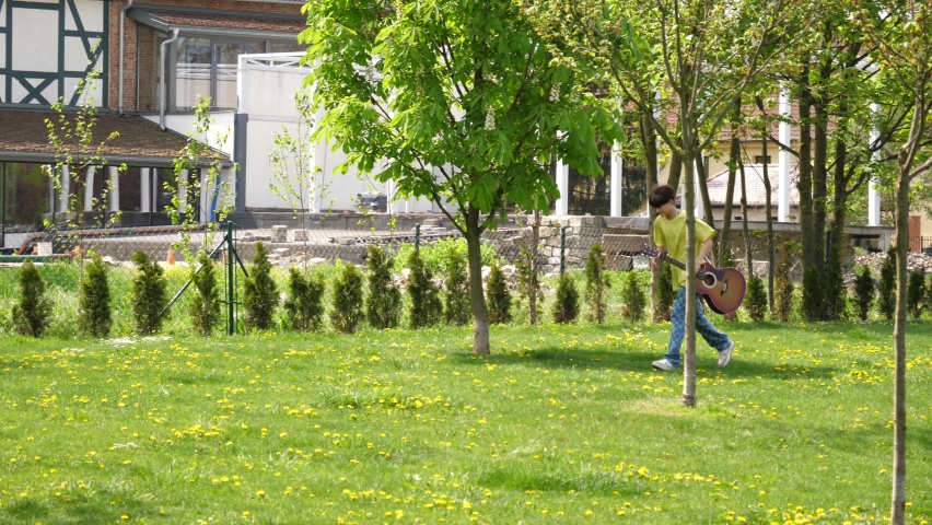 Teenager boy with guitar run through green park lawn hurrying somewhere