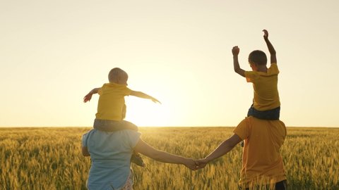 Parents in summer clothes carry sons in bright yellow t-shirts on shoulders and join hands walking on green field at sunset back view slow motion