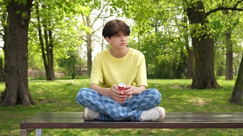 WROCLAW, POLAND - MAY 06, 2022: Teenager boy kid drinks cola beverage from a red can sitting on a park bench in a city