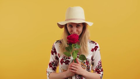 Studio shot of a pretty young cheerful woman with blonde hair in a fair hat and a floral blouse, standing and smelling a giant red rose. Valentine's concept. High quality 4k footage