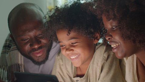 African American mother, father and little son lying together in dark room, smiling and watching something on digital tablet while spending evening at home