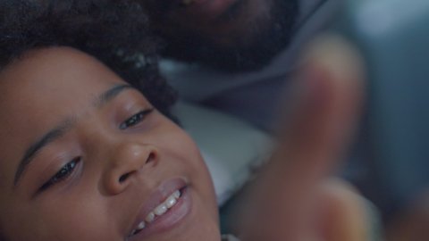Close up handheld camera shot of smiling African American father and little son lying together on bed and watching something on digital tablet