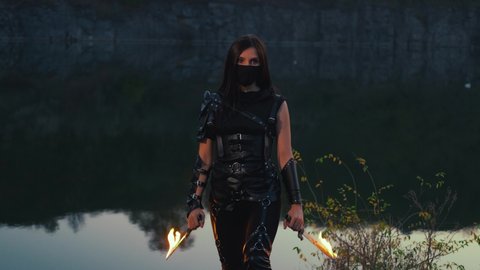 Beautiful fantasy woman warrior holding knives in hands. Fire swords burns brightly. Girl defender black leather ninja creative costume. face hide mask. Scenic night nature landscape rocky river bank 