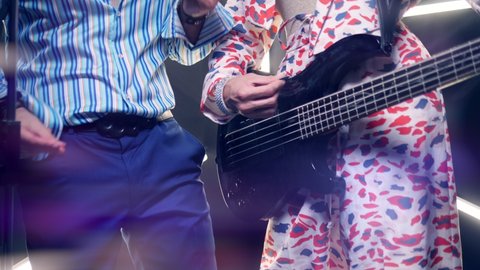 close-up of the hand. a girl in a bright suit plays the bass guitar, next to her a man in a shirt dances.