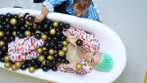 top view, rotating image. a travesty actor lies in a bathroom with gold and black balls, a happy man sits next to her on the floor.