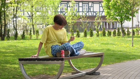 WROCLAW, POLAND - MAY 06, 2022: Kid boy teenager alone sit on a bench drinking coca cola can carbonated sweet beverage
